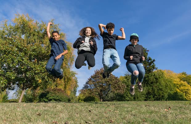 4 smiling teenagers shown jumping in midair on a sunny day in the park